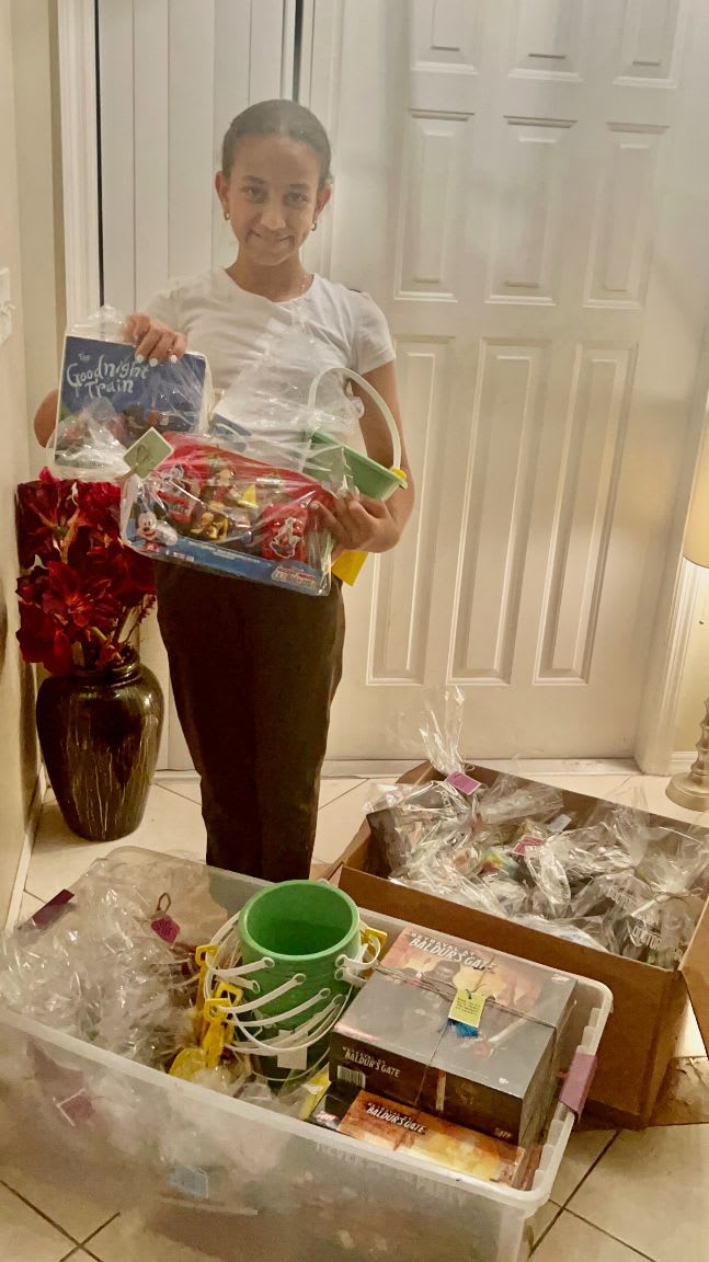 A young girl stands in a hallway holding a book and a basket of gifts, with several large bins filled with donations in front of her and a white door in the background.