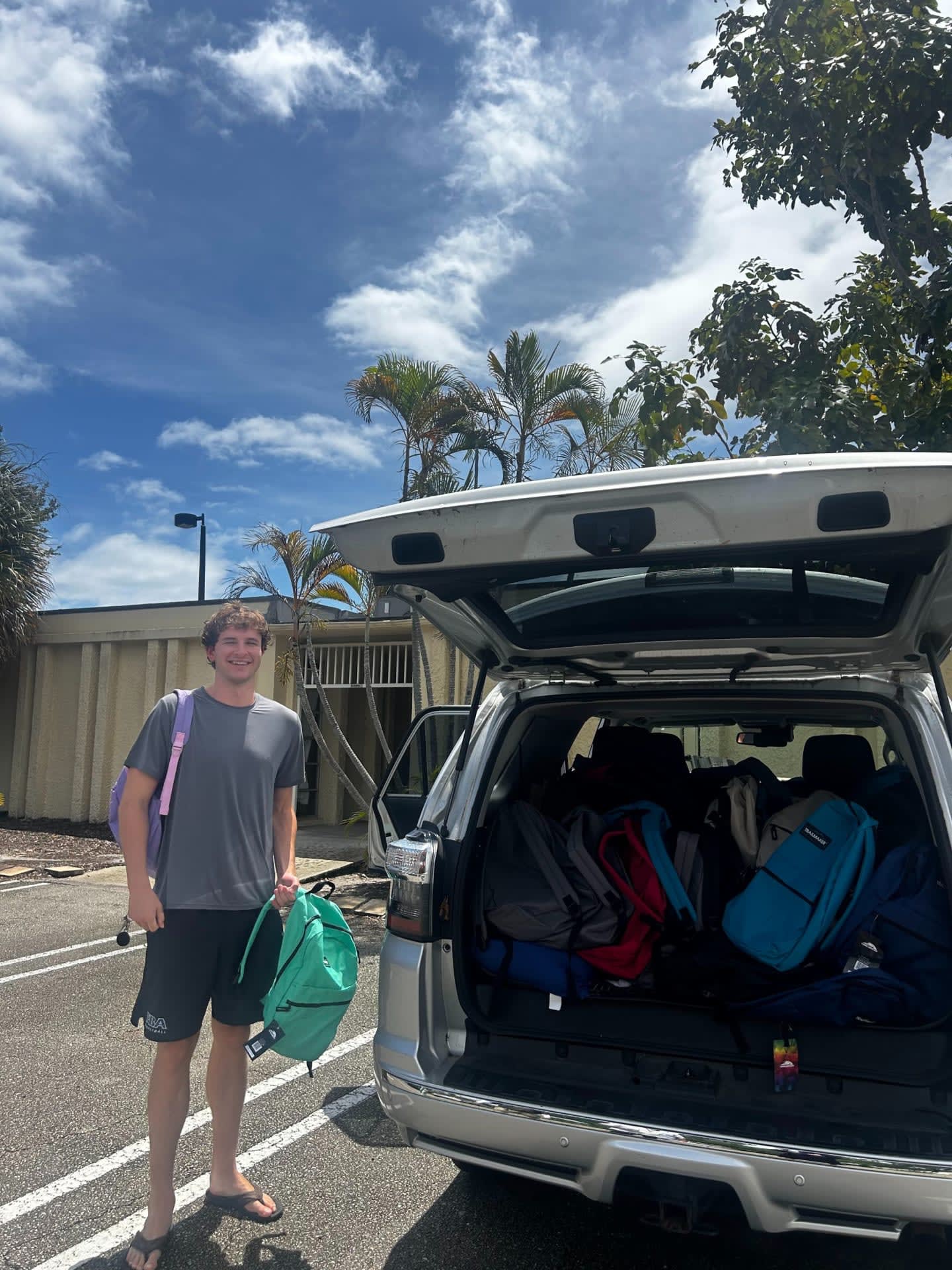 A young man stands by the open trunk of an SUV filled with backpacks, with palm trees and a building visible in the background under a cloudy sky.