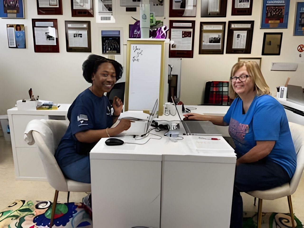 Two women smile at the camera from behind a white desk with laptops, while framed awards and certificates adorn the wall in the background.