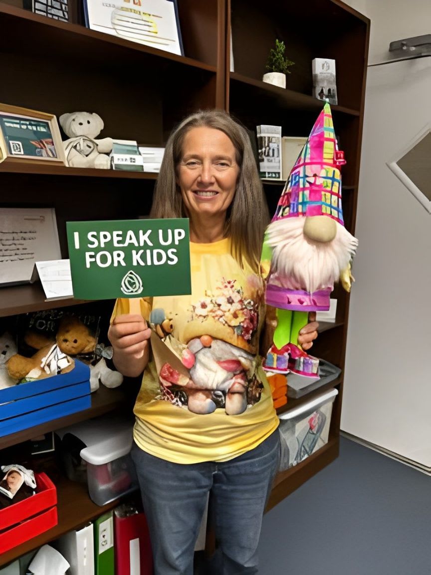 A woman in a gnome-themed shirt holds a sign that says "I SPEAK UP FOR KIDS" while standing in front of shelves filled with books and stuffed animals.