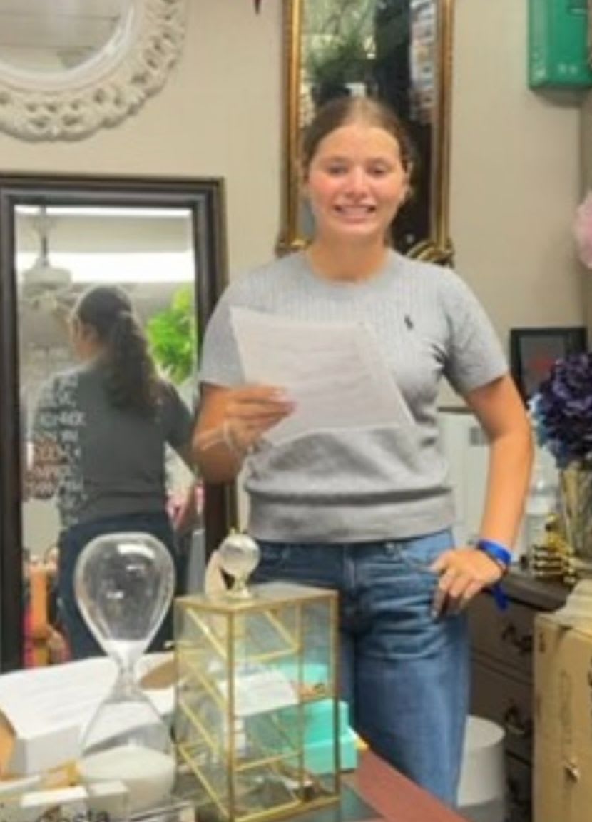 A smiling girl in a gray shirt and blue jeans stands in the foreground holding a paper, with a mirror reflecting another person and decorative items in the background.