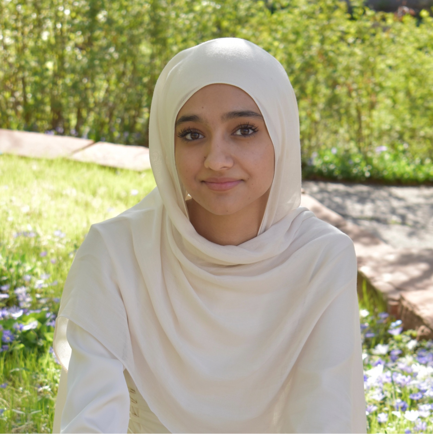 A young woman wearing a white hijab smiles gently in the foreground, with a blurred green and brown outdoor setting in the background.