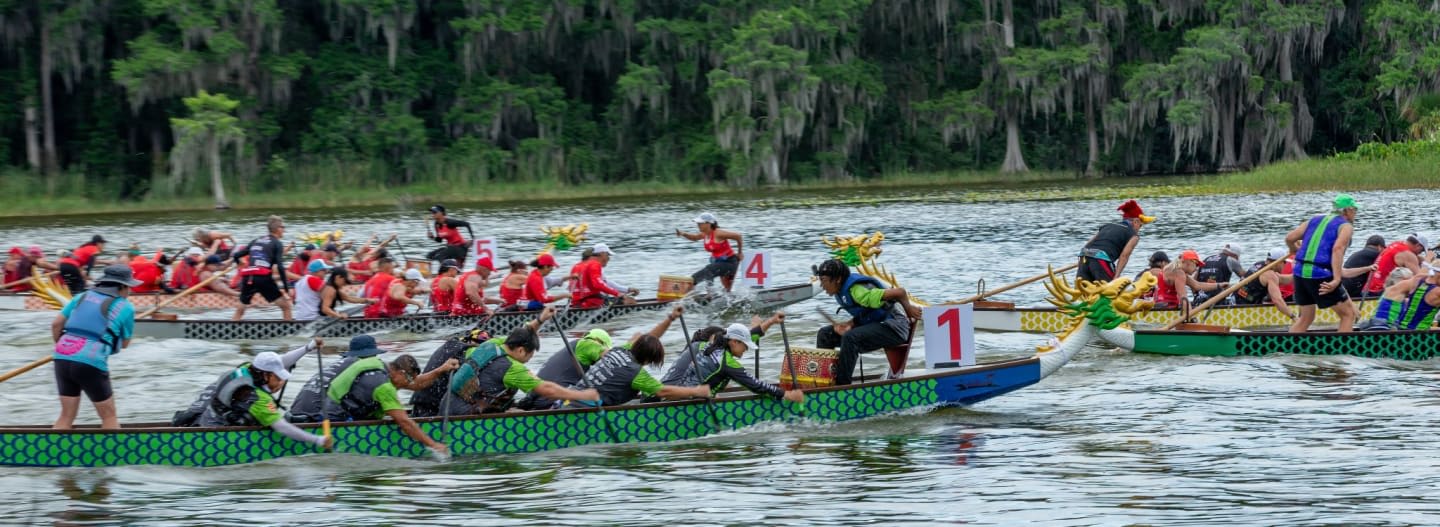Dragon boats race on a lake with a lush, tree-lined shore in the background.