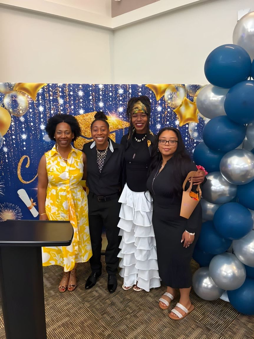 Four people stand in front of a graduation-themed backdrop with blue and gold balloons, while a black podium is visible in the foreground.