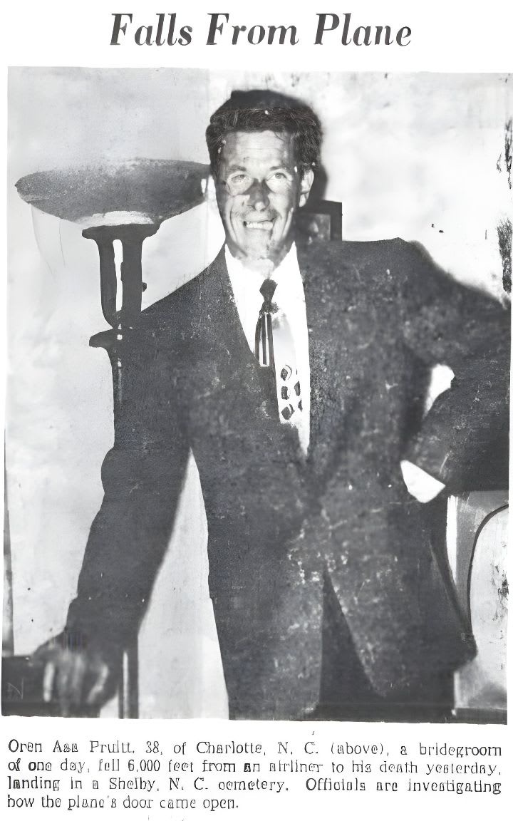 A smiling man in a suit stands in the foreground, with a lamp and a chair visible in the blurred background.