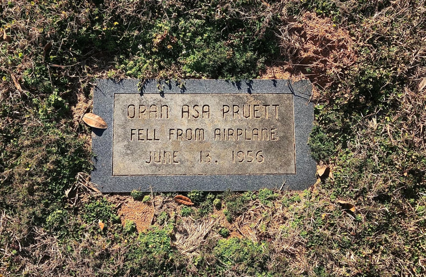 A gravestone with the inscription "OPAN ASA PRUETT FELL FROM AIRPLANE JUNE 13, 1956" is surrounded by dry grass and fallen leaves.