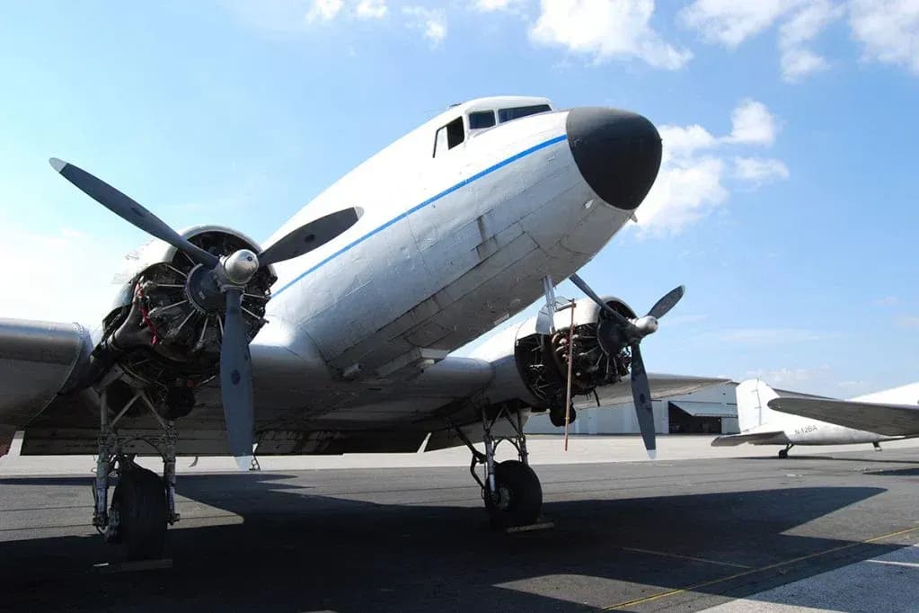 A vintage white airplane with blue trim sits on an asphalt tarmac under a bright blue sky with scattered clouds, with another aircraft visible in the background.