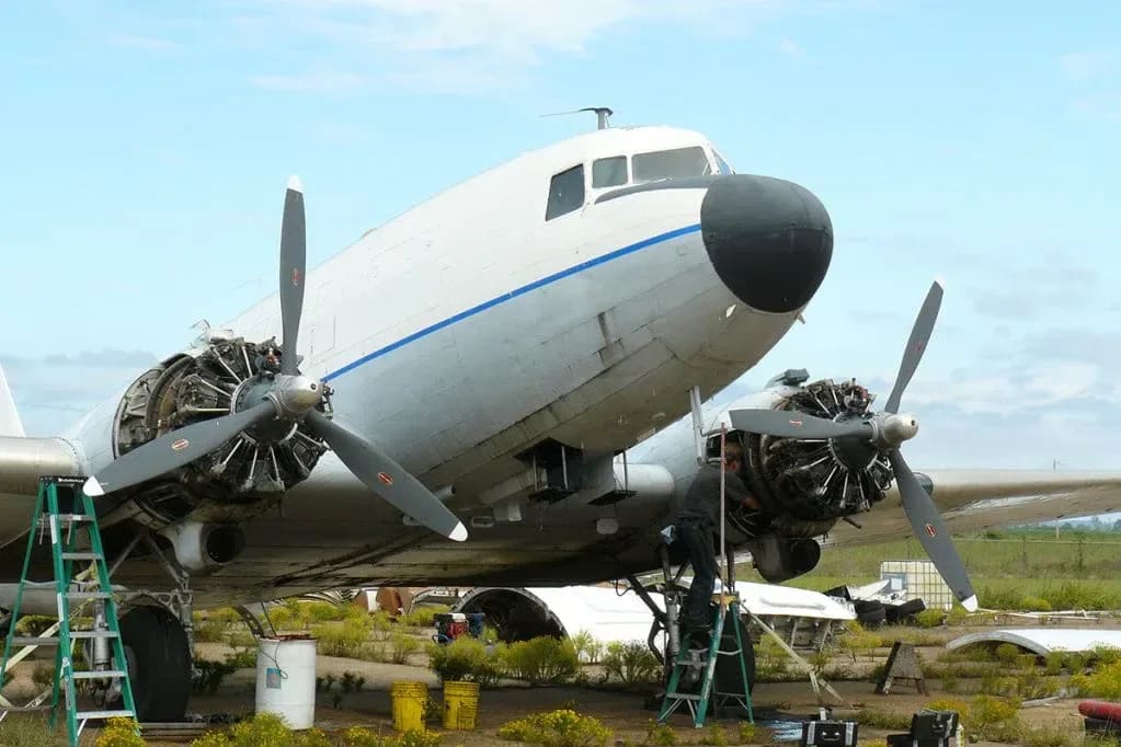 A vintage white airplane with exposed engines sits in a grassy field under a cloudy sky, with ladders and equipment around its fuselage.