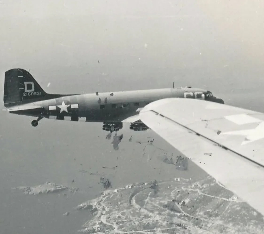 A military transport plane flies over a coastal landscape, with a wing of another aircraft in the foreground.