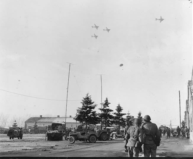 Soldiers stand in the foreground watching planes fly overhead, while military vehicles and buildings are visible in the background.