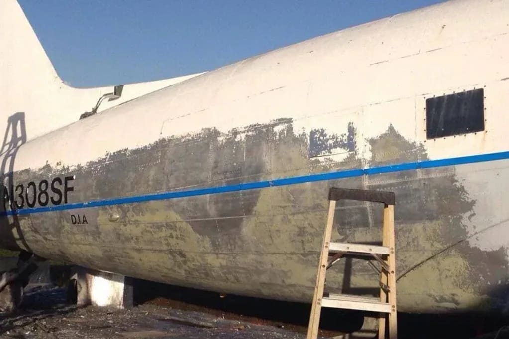A wooden ladder leans against the partially stripped fuselage of a white airplane with blue stripes, set against a clear blue sky.