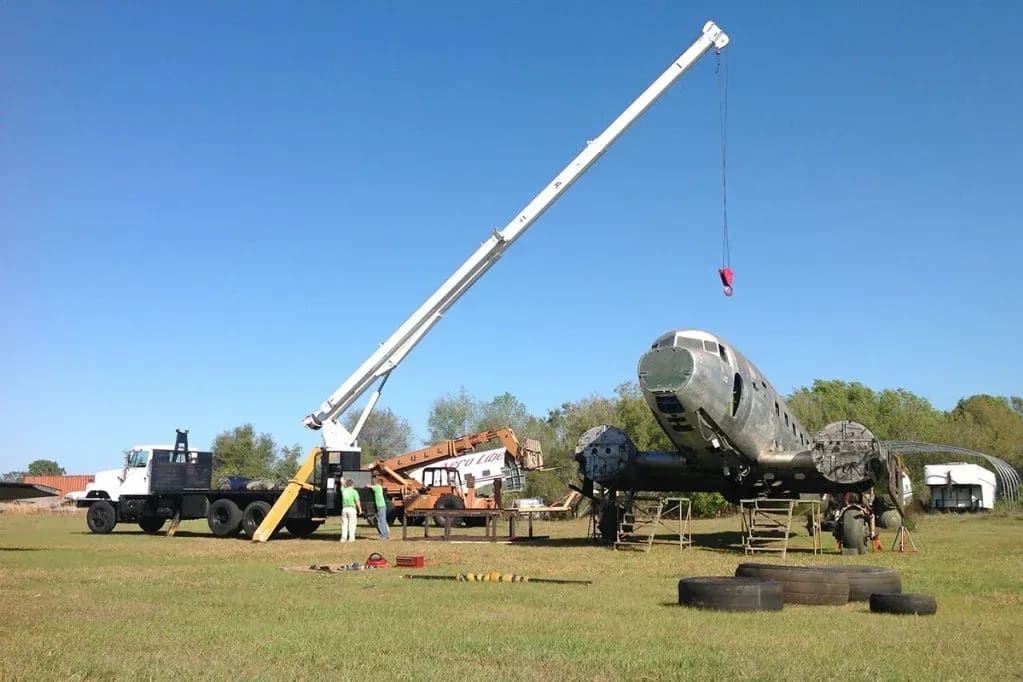 A large crane lifts a section of an old airplane in a grassy field with trees and buildings in the background.