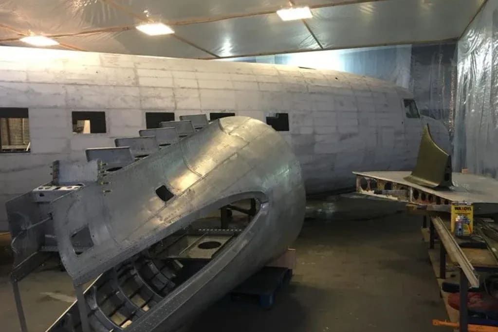 A partially disassembled aluminum airplane fuselage sits in a workshop with a large, unfinished aircraft in the background.
