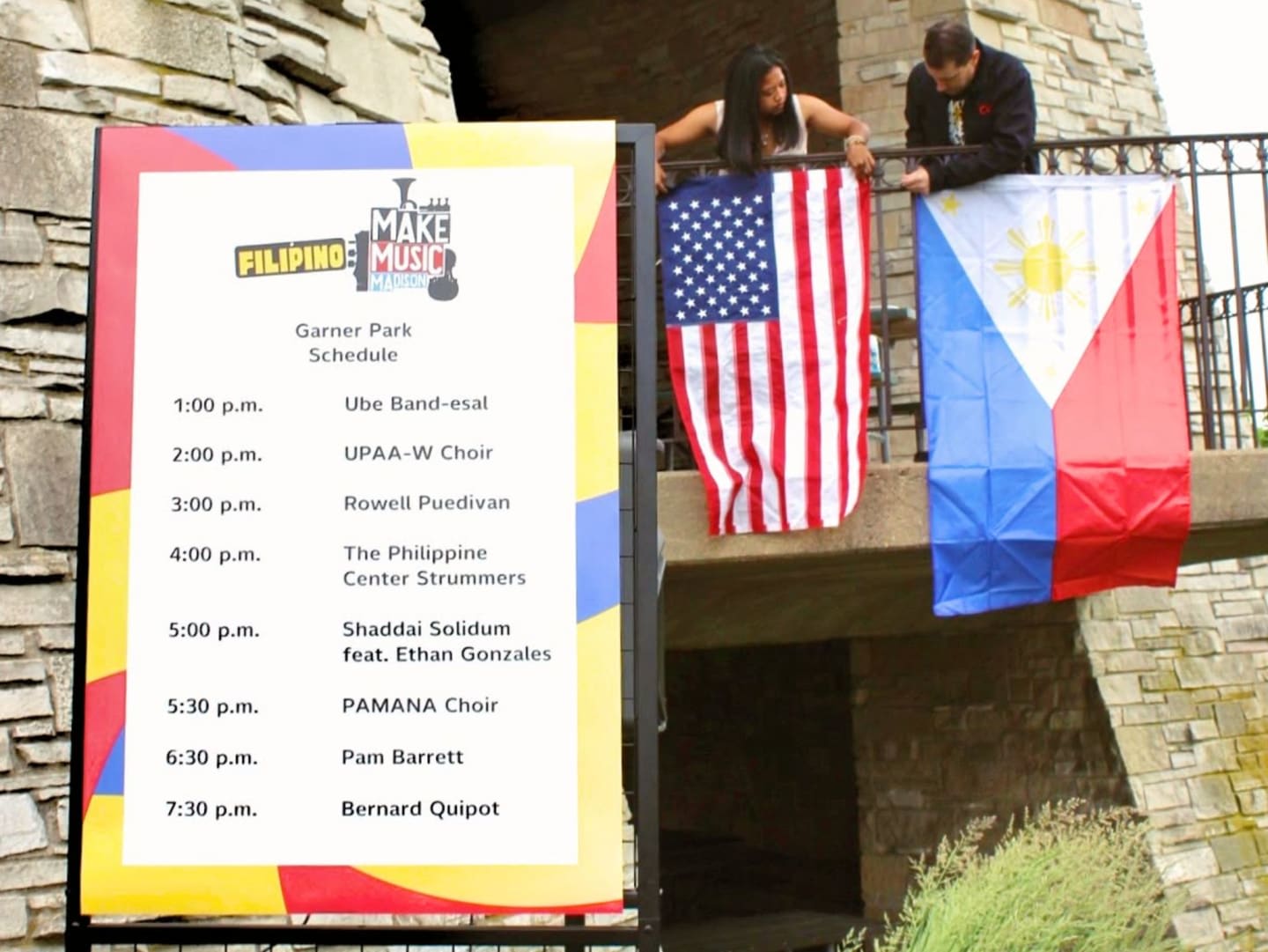 A colorful schedule for Filipino Music at Garner Park stands in the foreground, while two people hang American and Philippine flags in the background.