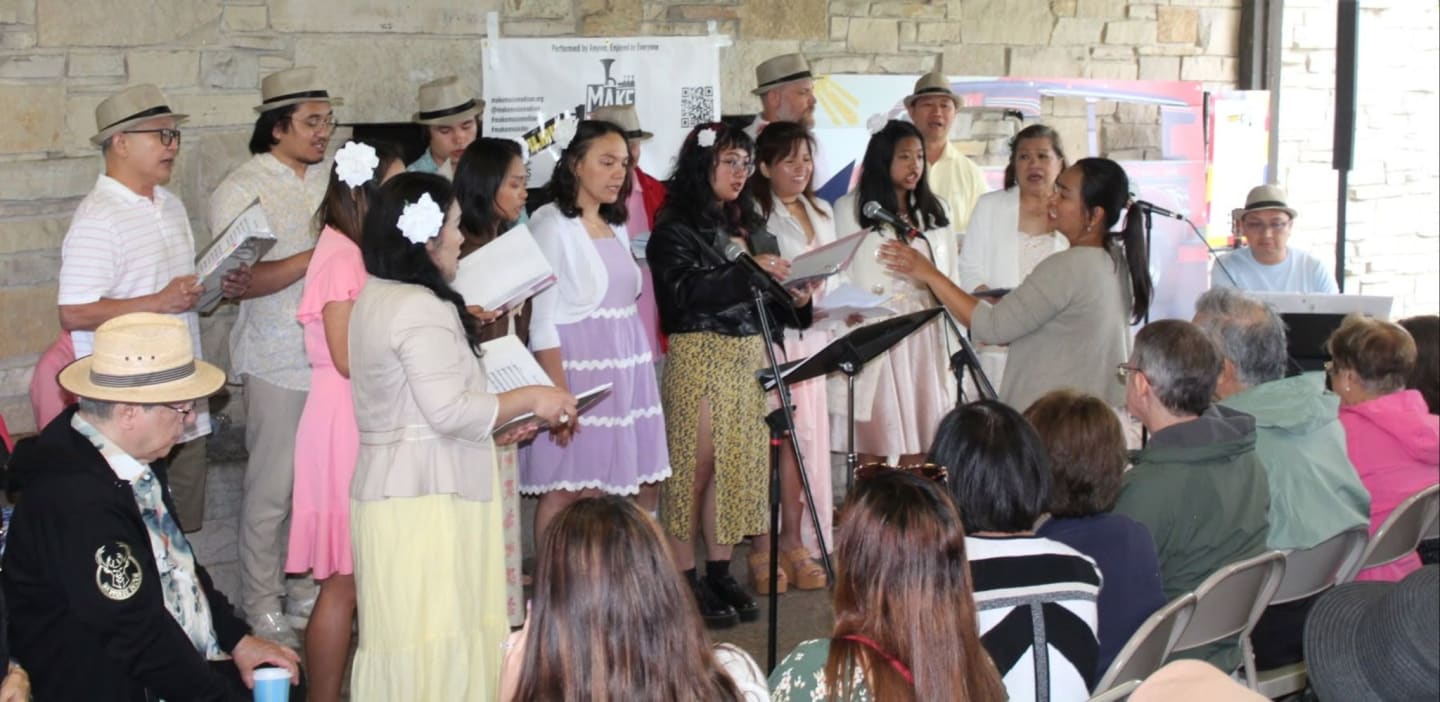 A choir of people in vintage attire sings in front of a stone wall, while an audience watches from chairs in the background.