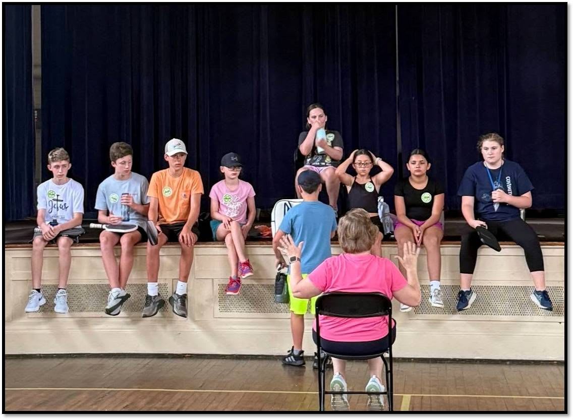 A group of children sit on a raised platform in front of a blue curtain, while an adult in a pink shirt faces them from a chair on the floor.