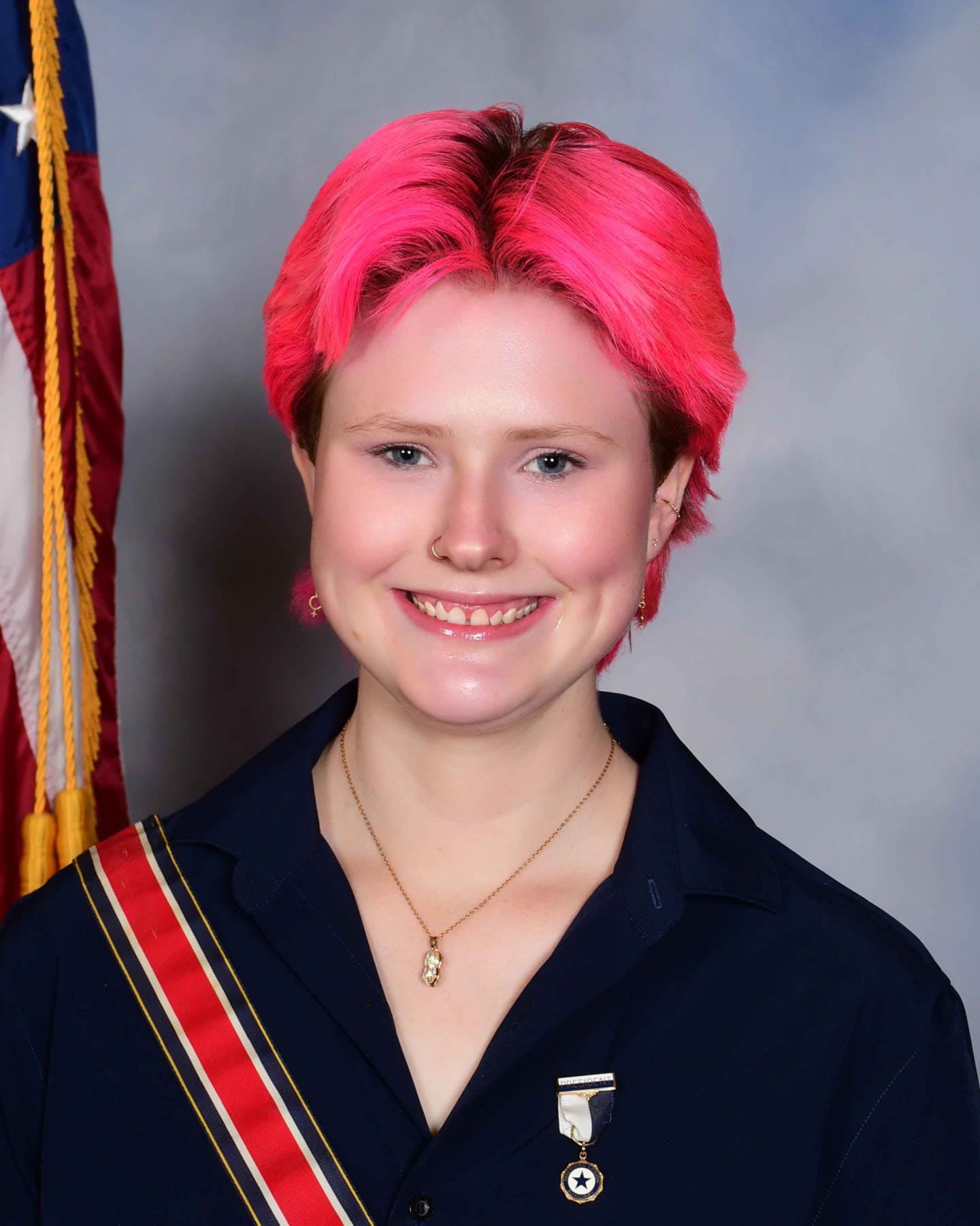 A young person with bright pink hair smiles in the foreground, with an American flag and a gray backdrop behind them.
