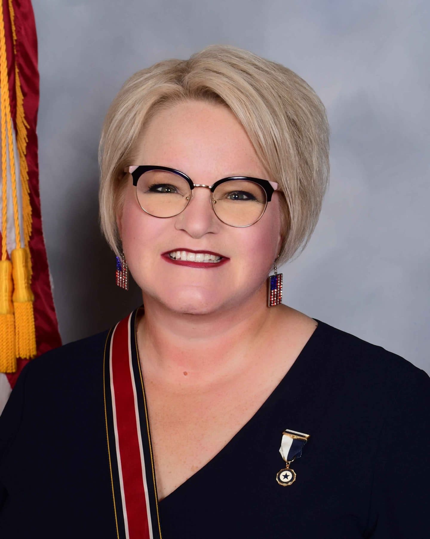 A woman with blonde hair and glasses smiles at the camera, wearing a dark blue dress with a sash, while a flag is visible in the background.