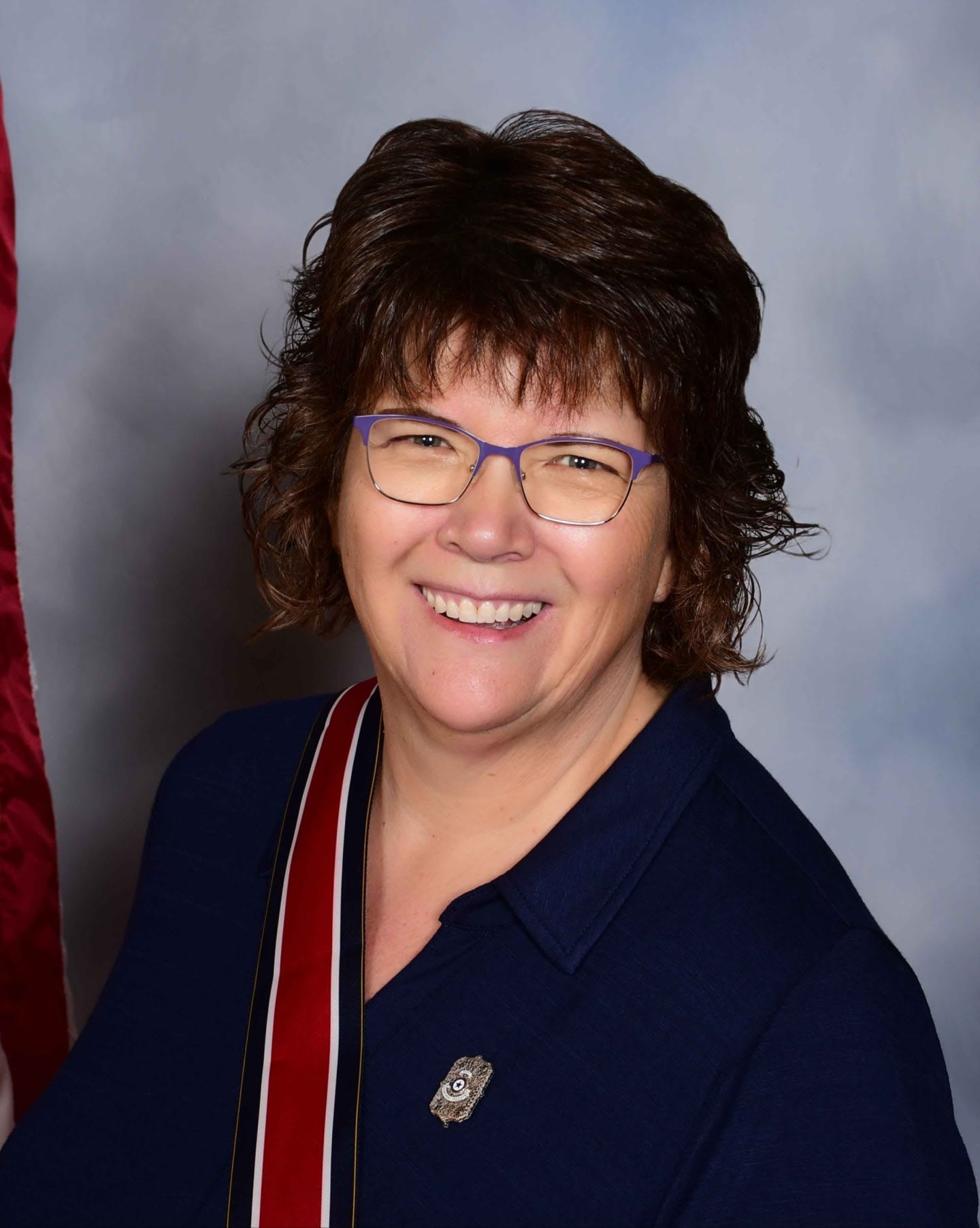 A smiling woman with glasses and a dark blue shirt stands in front of a flag and a mottled blue background.