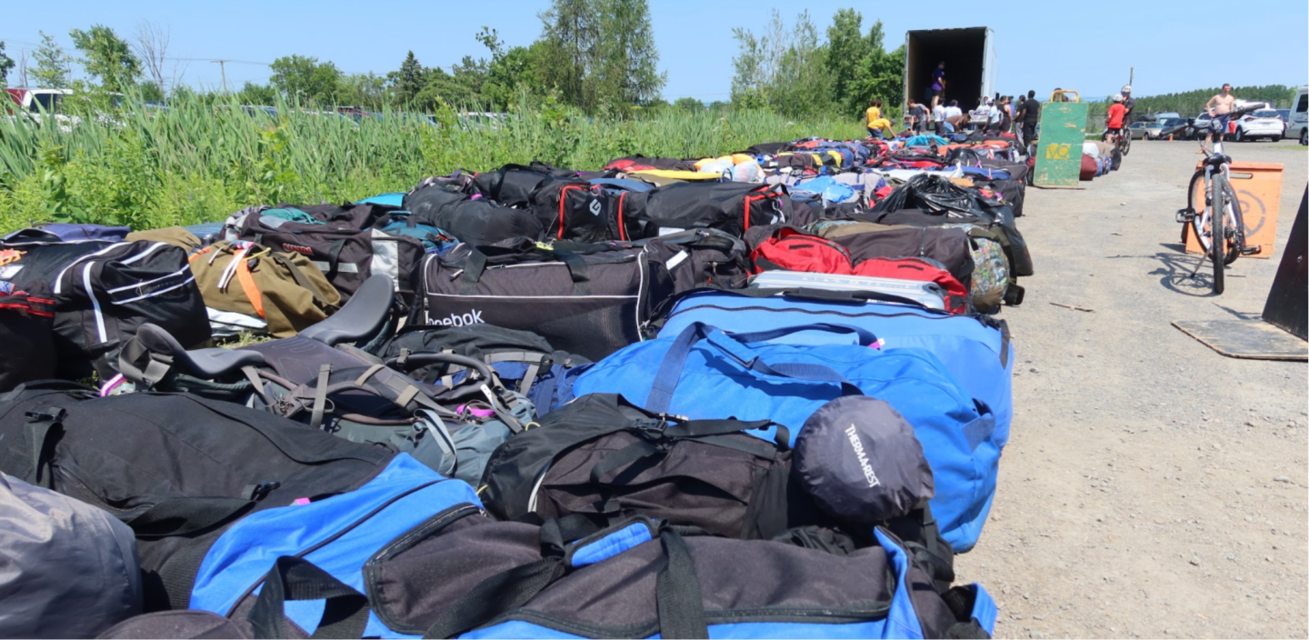 A large collection of duffel bags is spread across a dirt lot in the foreground, with a truck and people in the background.