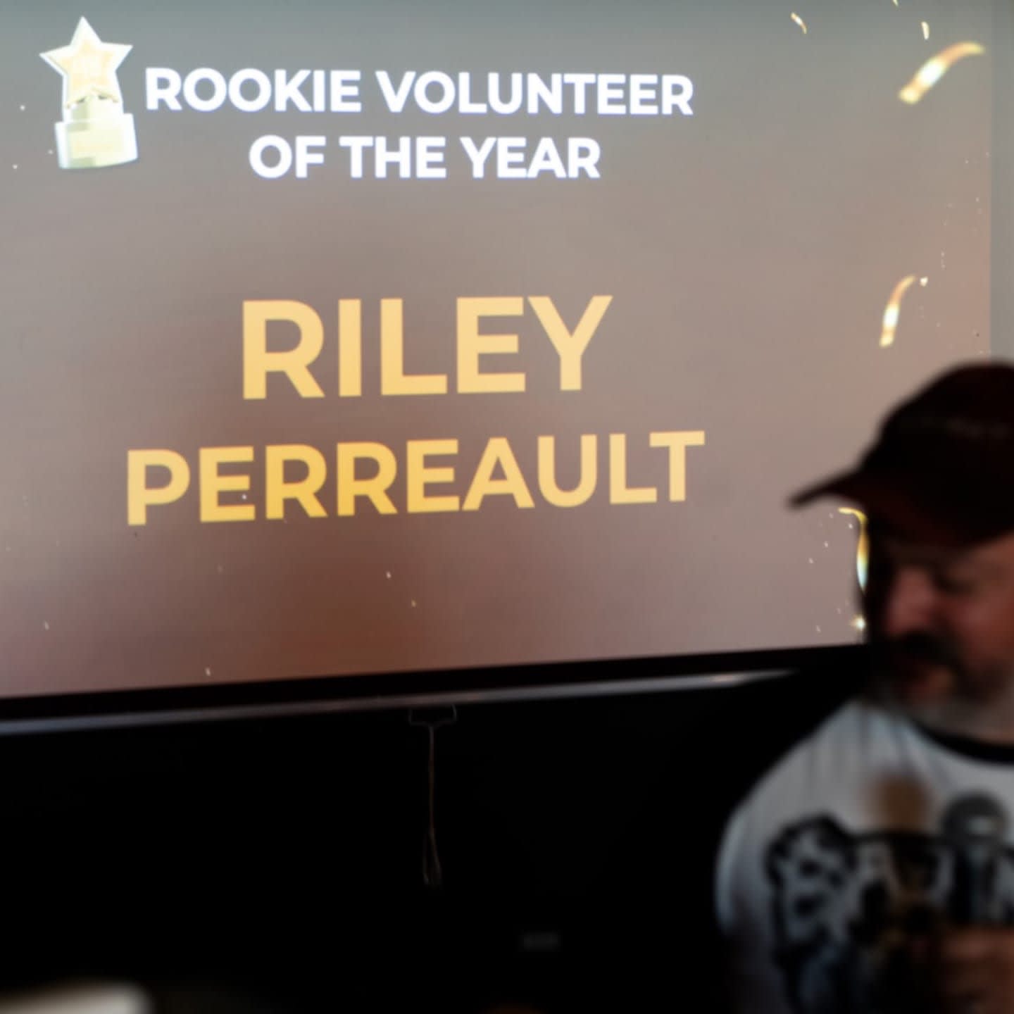 A man in a baseball cap and t-shirt stands in the foreground, partially obscuring a screen in the background that displays "ROOKIE VOLUNTEER OF THE YEAR RILEY PERREAULT" with confetti.