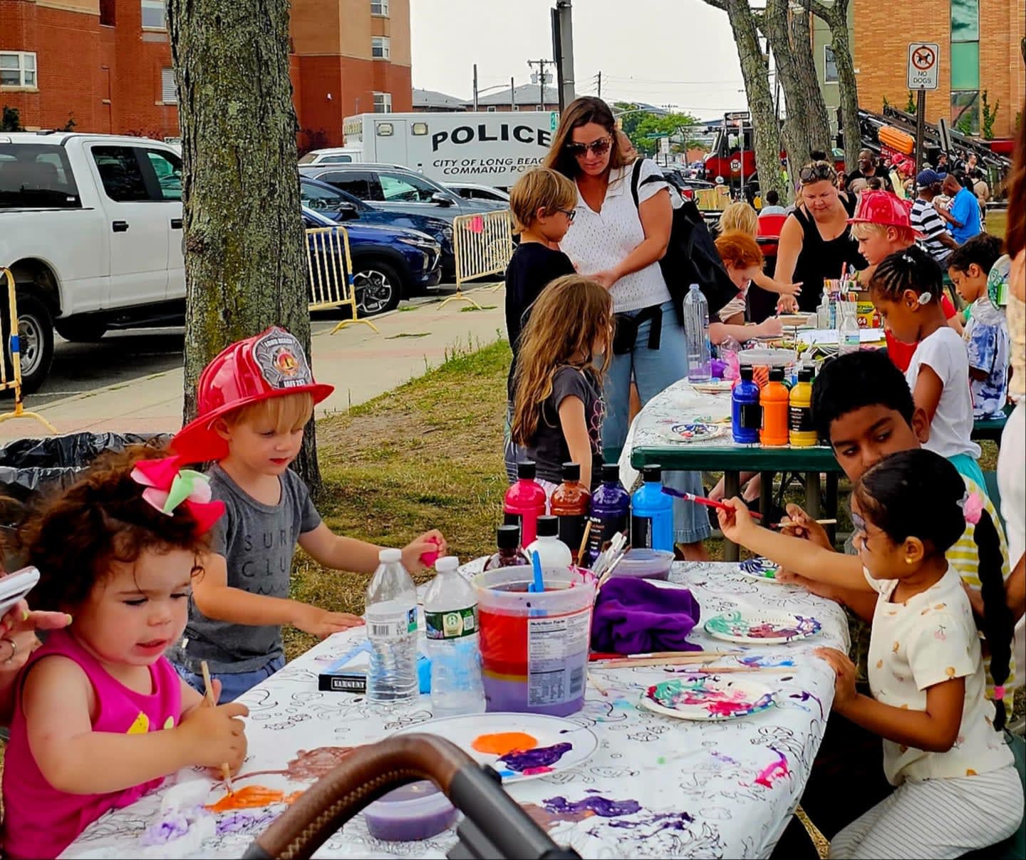 Children in the foreground are engaged in a painting activity at tables, while in the background, adults and a police vehicle are visible.