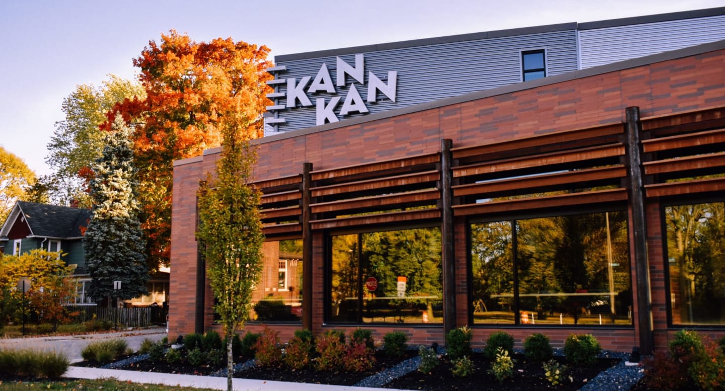 A modern brick building with large windows and a sign reading "EKAN KAN" stands in the foreground, with colorful autumn trees and a house visible in the background.