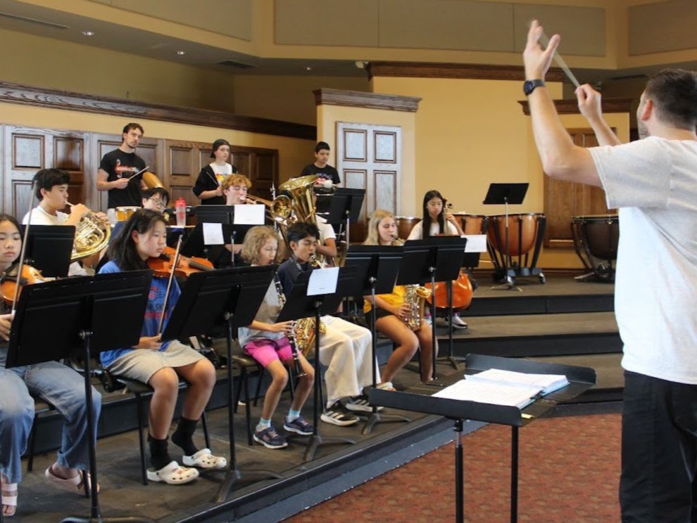 A conductor leads a youth orchestra in a rehearsal, with students playing various instruments in the foreground and a warm, wood-paneled room in the background.