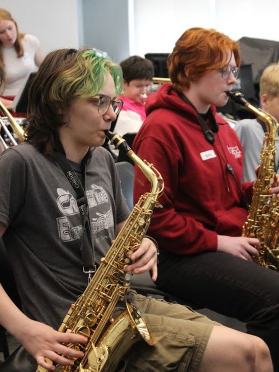 Two young people with saxophones in the foreground are playing in a band with other musicians in the blurred background.