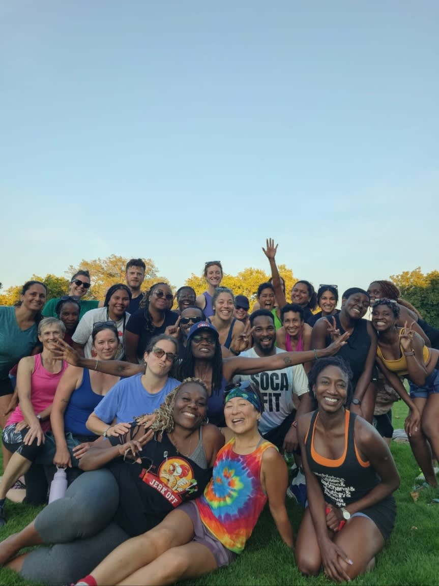 A diverse group of people in athletic wear pose for a photo on a grassy field with a clear sky in the background.