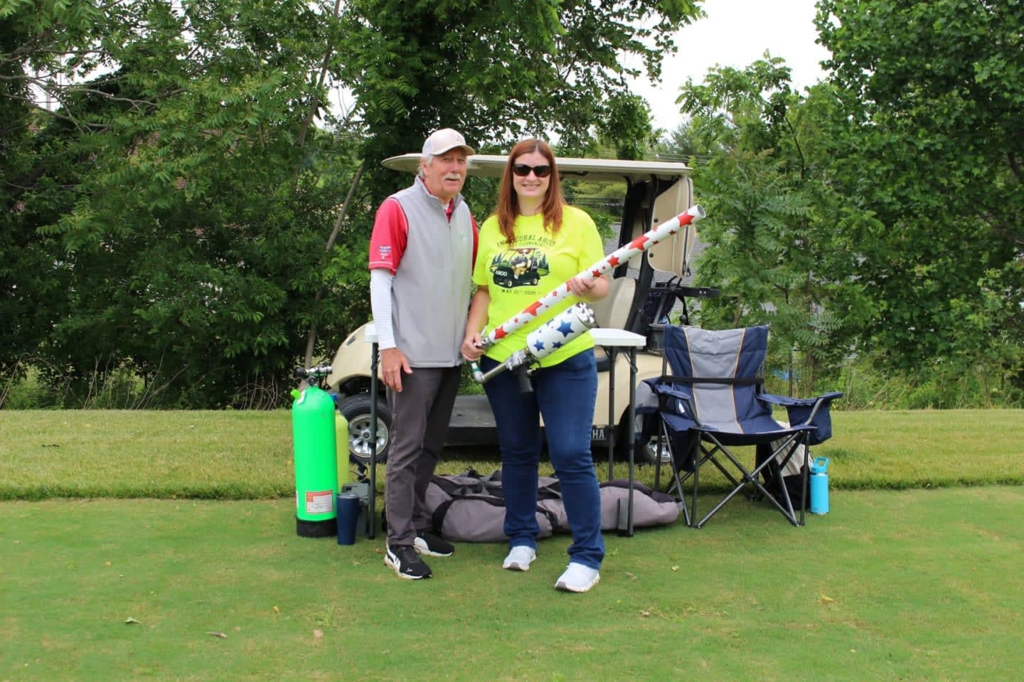 A man and woman stand on a golf course in front of a golf cart and chairs, with trees in the background.