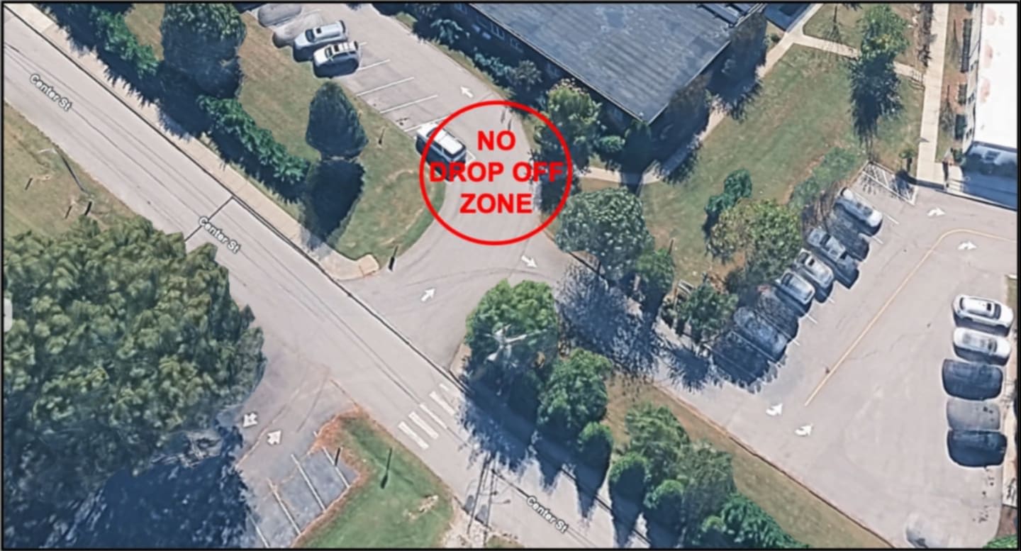 A white car is parked within a red circle marked "NO DROP OFF ZONE" in the foreground, while a parking lot filled with cars and a building are visible in the background.