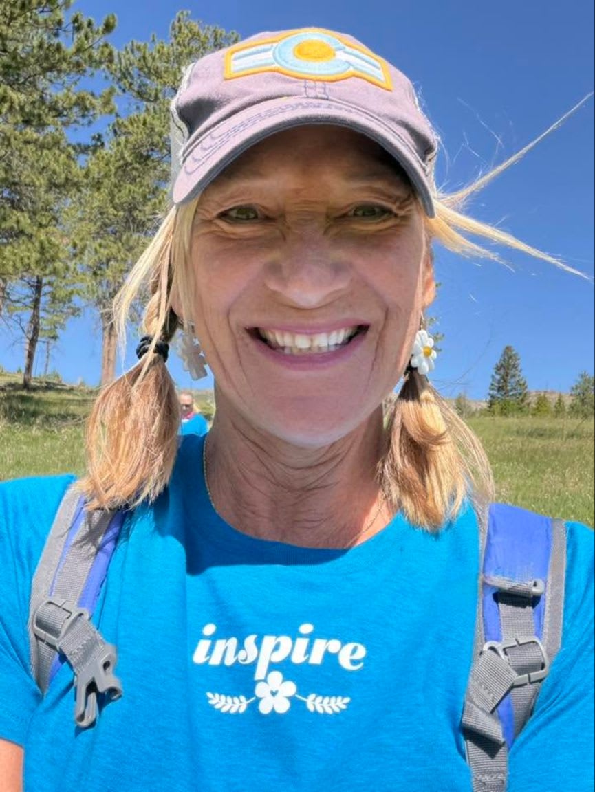 A smiling woman with blonde pigtails and a blue "inspire" t-shirt is in the foreground, with a grassy, tree-dotted hillside and a bright blue sky in the background.