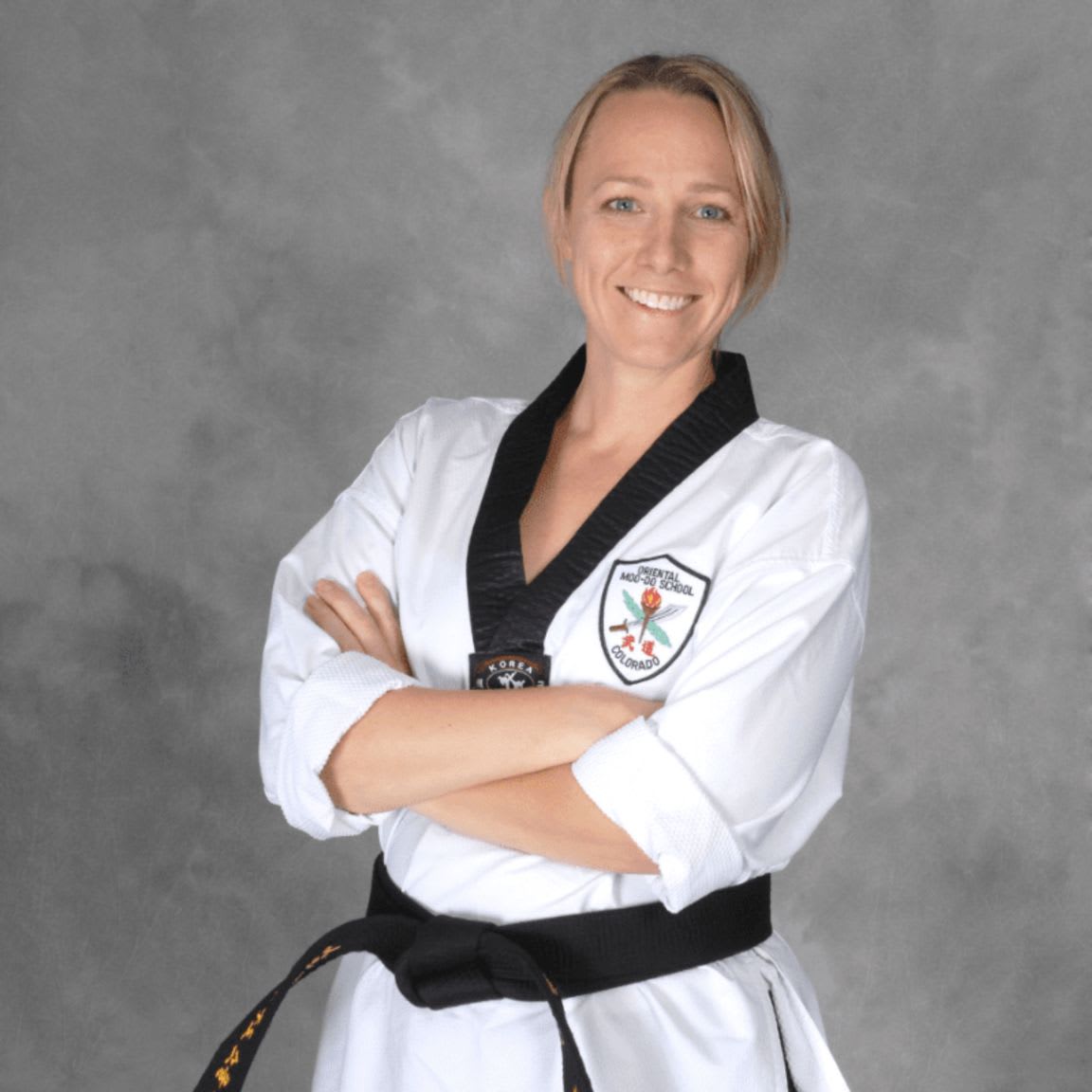 A smiling woman in a taekwondo uniform with her arms crossed stands against a textured gray background.