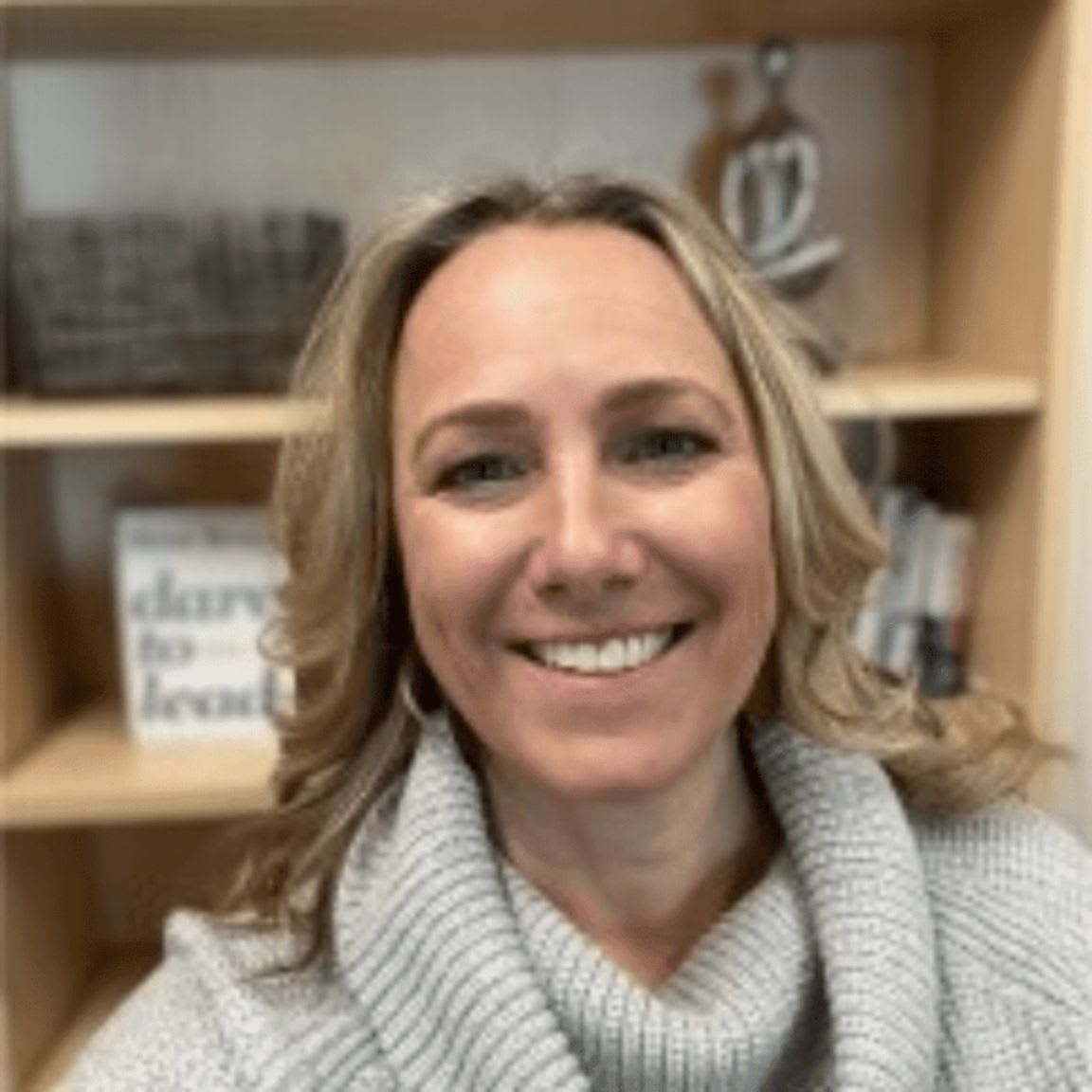 A smiling woman in a gray sweater is in the foreground, with a bookshelf containing books and decorative items in the background.