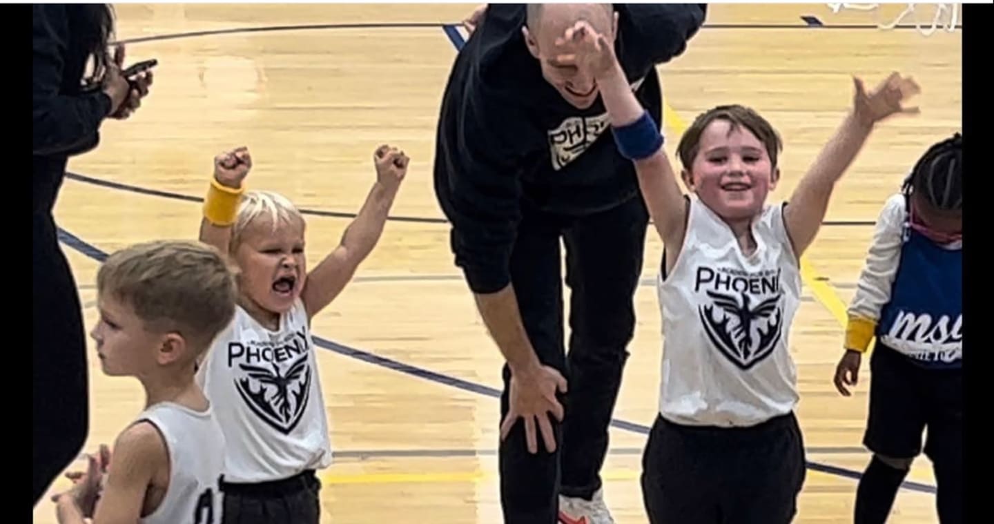 Young boys in basketball jerseys cheer with raised arms on a wooden court, while an adult watches them in the background.