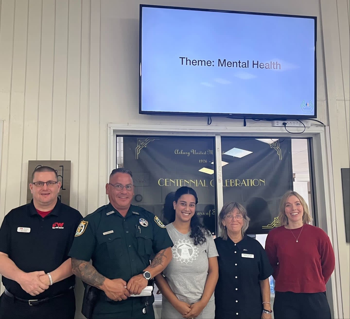 Five people stand smiling in the foreground in front of a door and a large screen displaying the text "Theme: Mental Health."