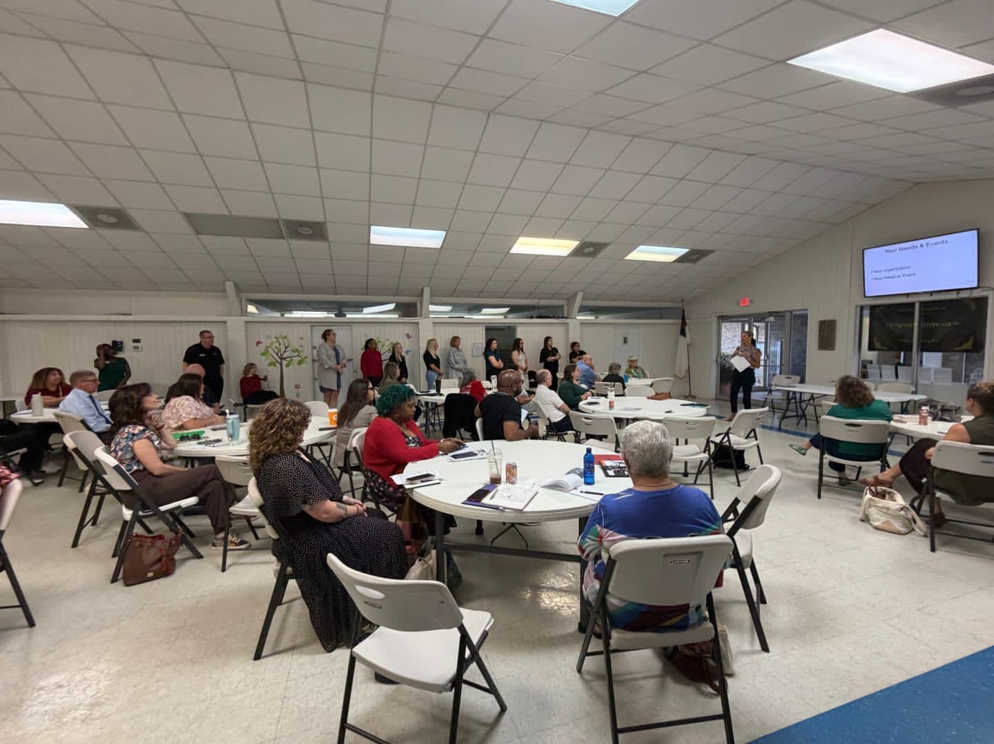 People sit at round tables in the foreground of a brightly lit community room, while others stand and converse in the background.