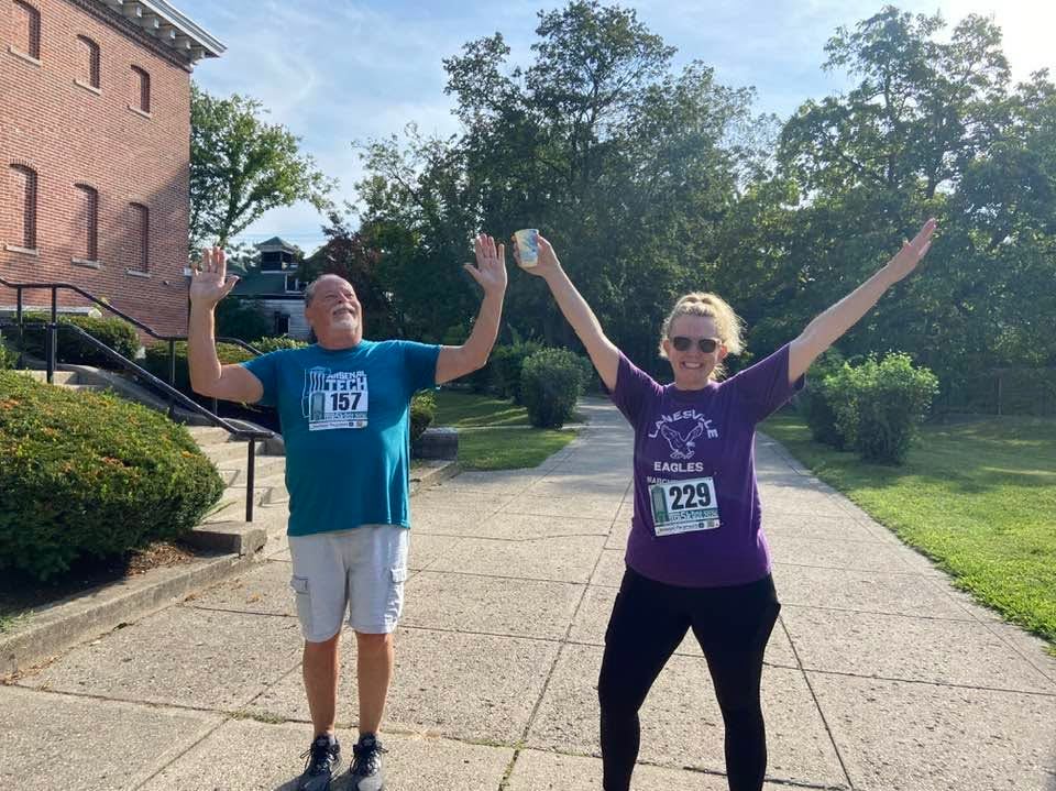 Two smiling runners with race bibs stand with their arms raised on a paved path, with a brick building and lush green trees in the background.