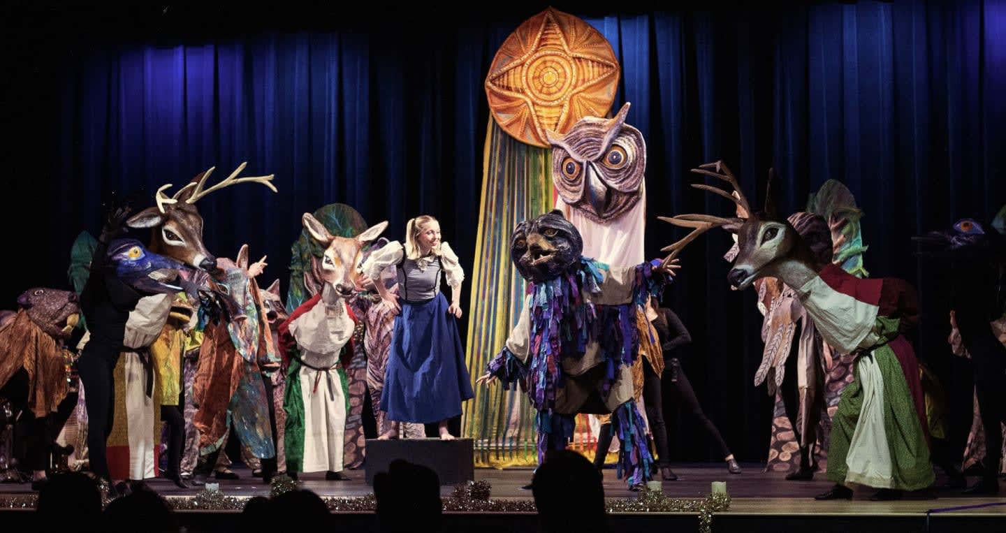 A woman in a blue dress stands center stage surrounded by performers in elaborate animal costumes against a backdrop of blue curtains and a large golden sun ornament.