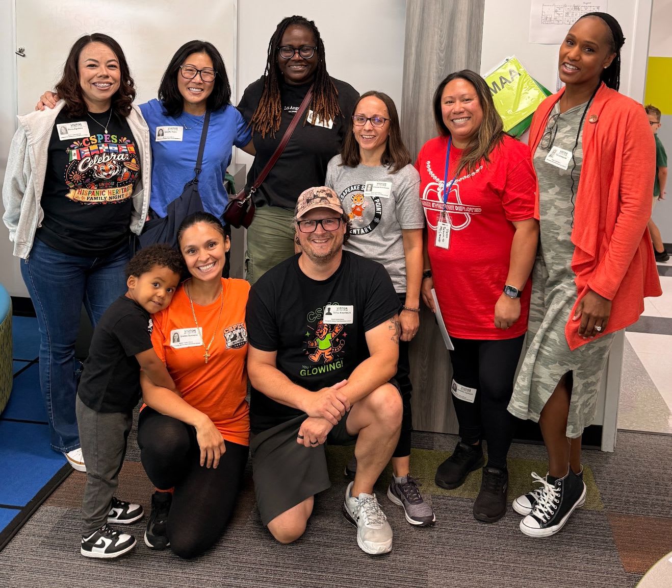 A diverse group of smiling adults and a young child pose together in a brightly lit indoor hallway.