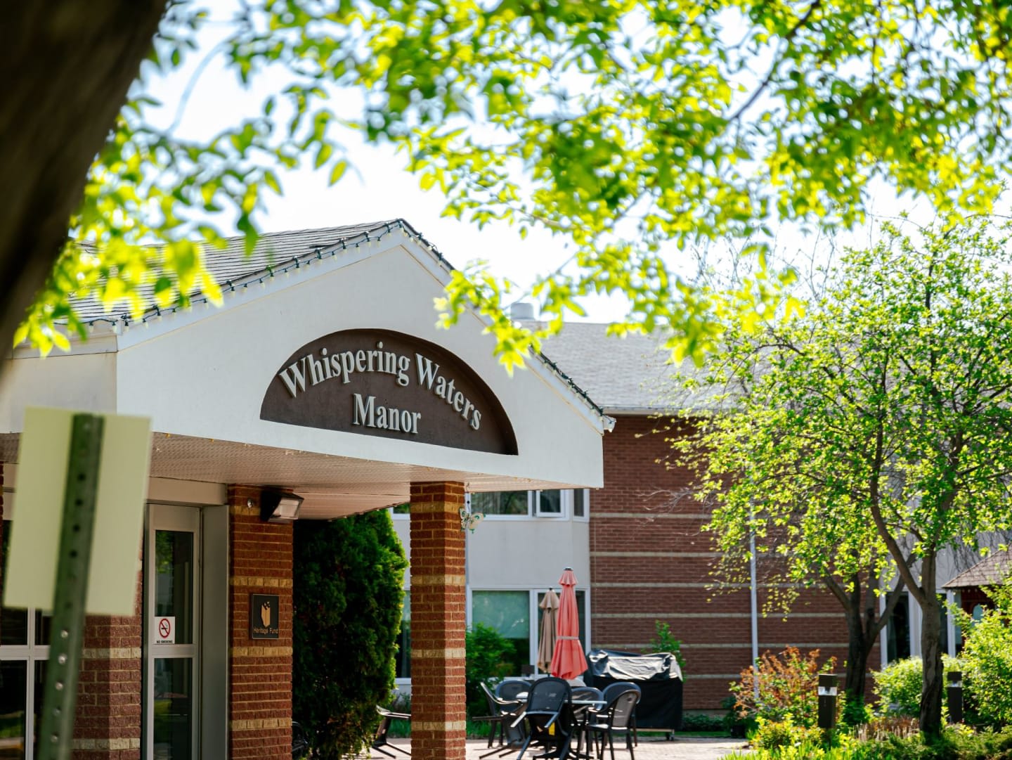 Green leaves frame the foreground of a brick building with a sign reading "Whispering Waters Manor" and a patio area in the background.