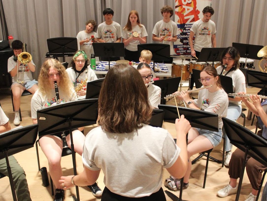A young conductor leads a student band consisting of wind and percussion players seated in a rehearsal room.