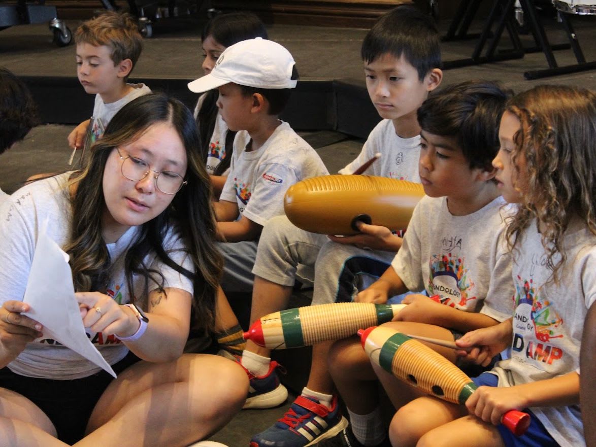 A woman reads a paper to a group of children holding wooden instruments in an indoor setting.