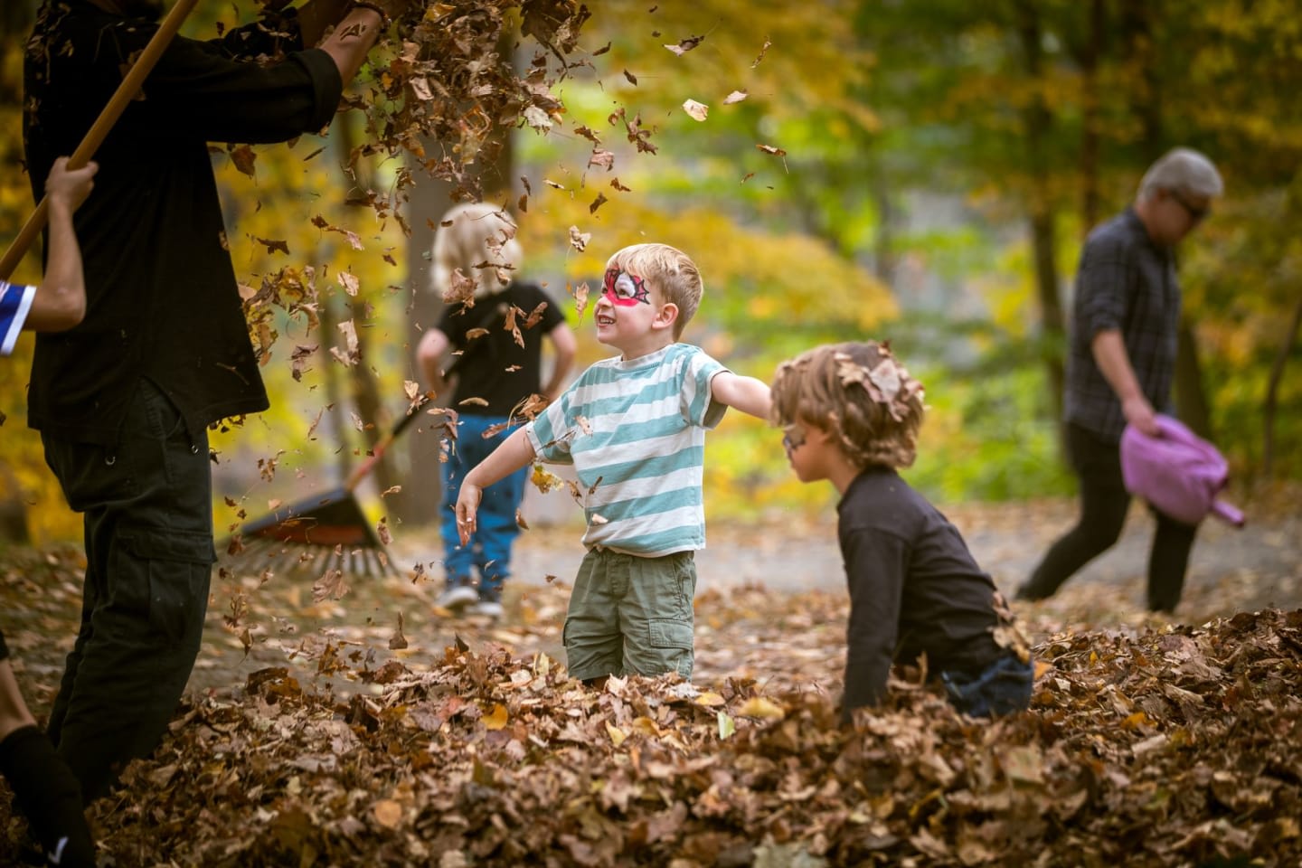 Children play in a large pile of autumn leaves in the foreground while a person rakes and a man walks in a wooded background.