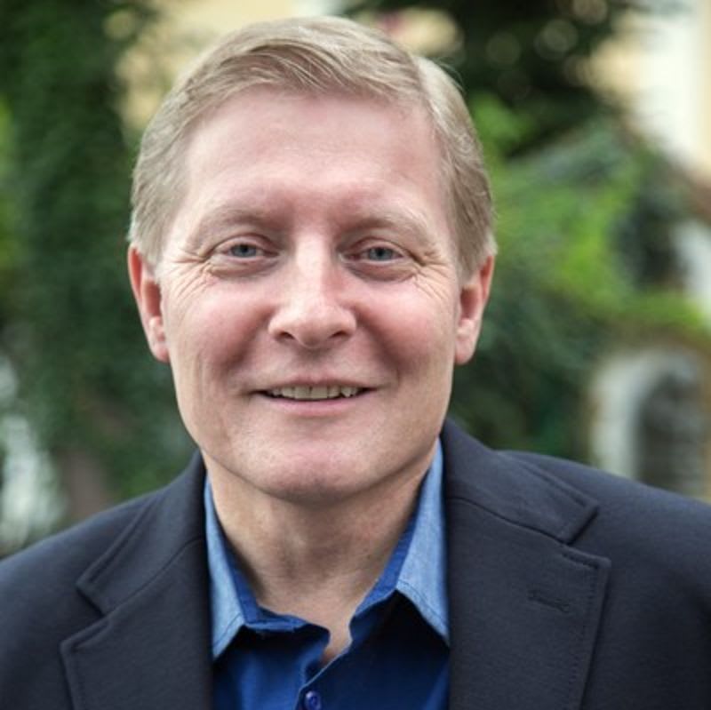 A smiling man in a dark blazer and blue shirt stands before a blurred background of green foliage.