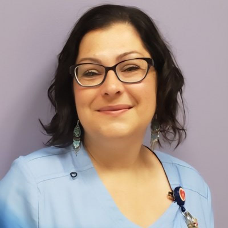 A woman wearing glasses and blue scrubs smiles in front of a plain purple wall.