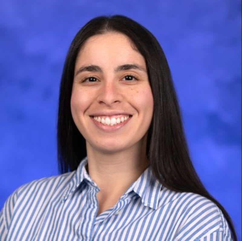 A smiling woman with long dark hair wearing a blue and white striped shirt stands against a solid blue background.