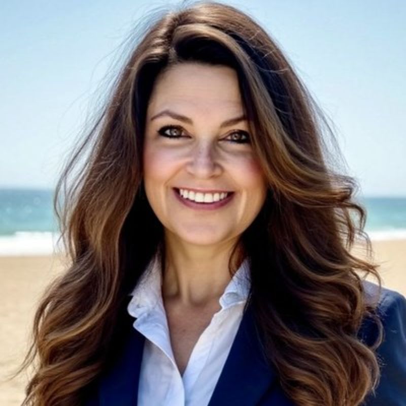 A smiling woman with long brown hair wearing a blue blazer and white shirt stands before a sandy beach and blue ocean.