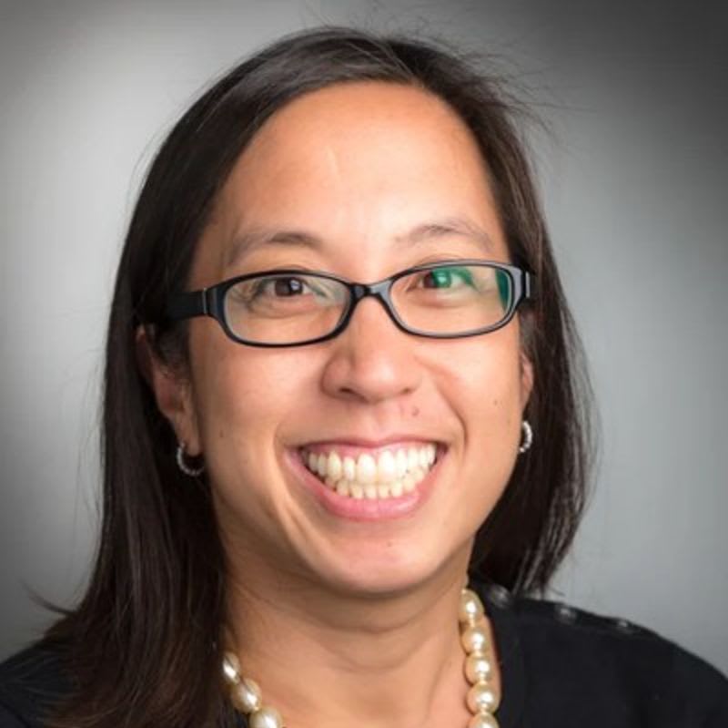 A smiling woman with glasses and a pearl necklace is positioned against a neutral gray background.